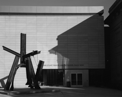 A large metal sculpture stands in front of a nearly blank wall. The wall has a grid pattern, and the shadow of a connecting older building.