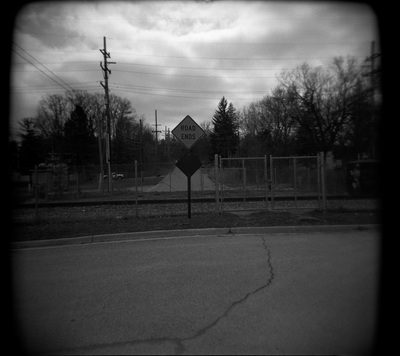 A sign blocks the road before it comes to a fence and a railroad. More road is visible on the other side of the tracks.